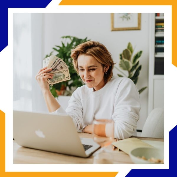 A smiling woman at a desk with a laptop holds a fan of cash, representing savings on energy bills.