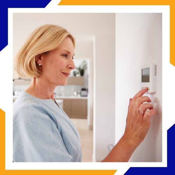 A middle-aged woman with blonde hair smiles as she adjusts the temperature on a digital wall thermostat.